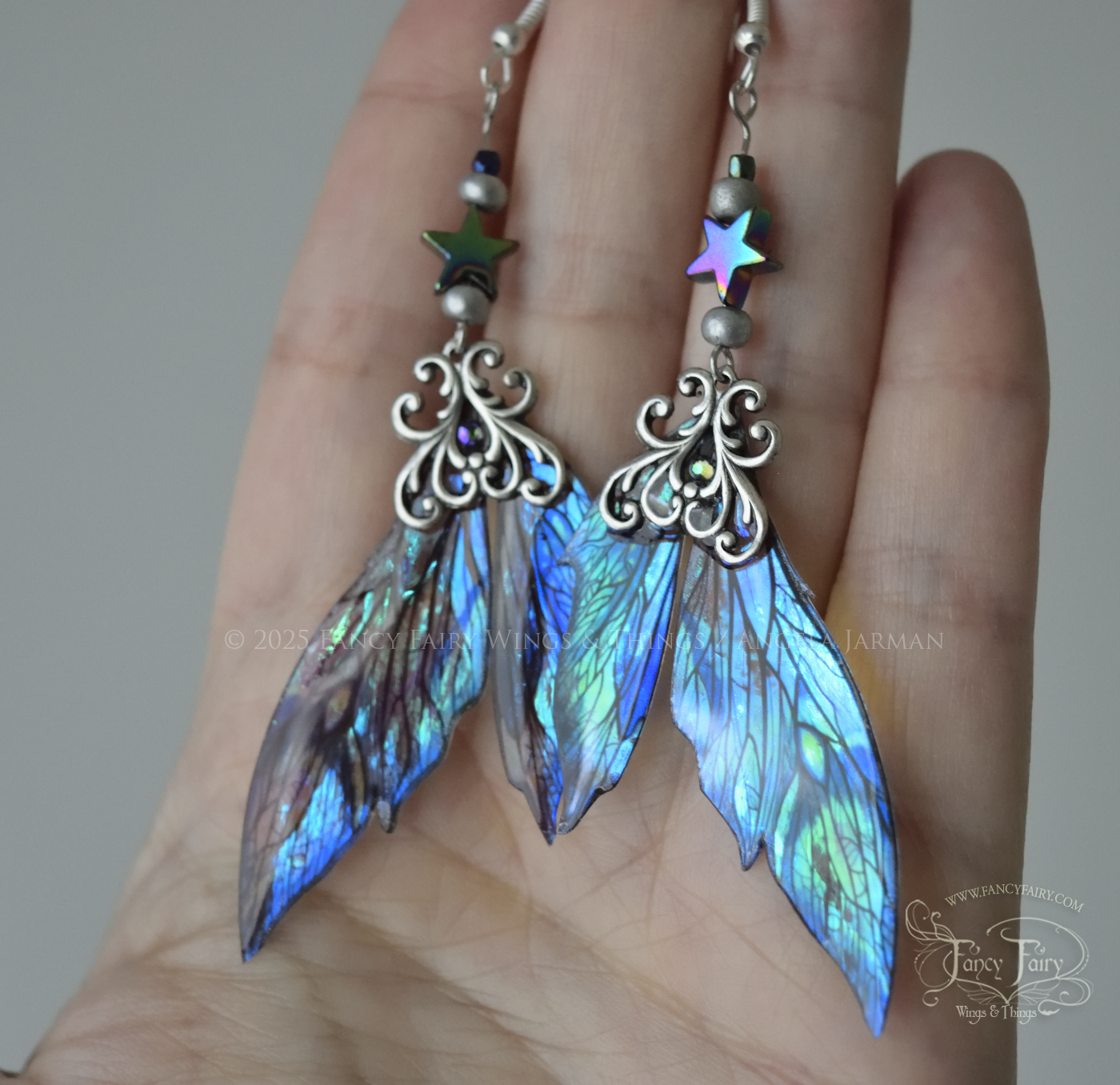 Iridescent fairy wing earrings in blues with silver filigree are held in the palm of a white woman's hand