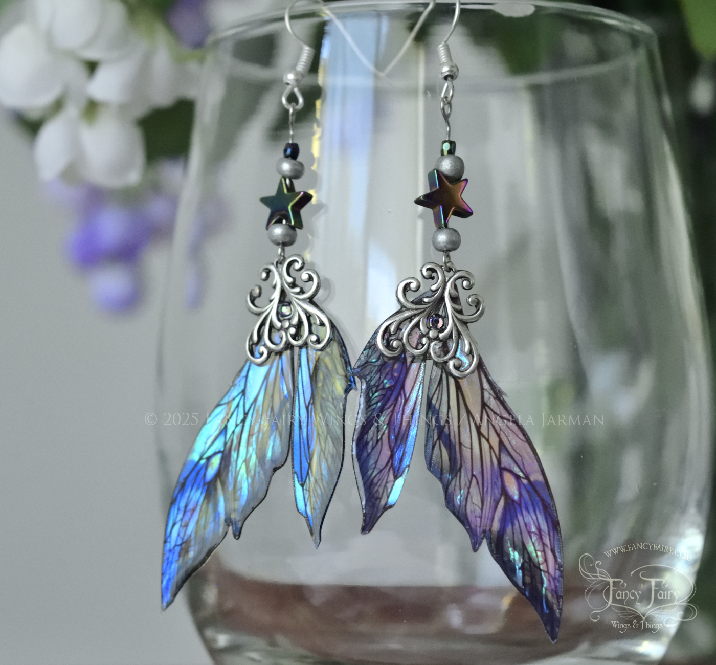 Iridescent fairy wing earrings in blues with silver filigree dangle from the rim of a clear glass