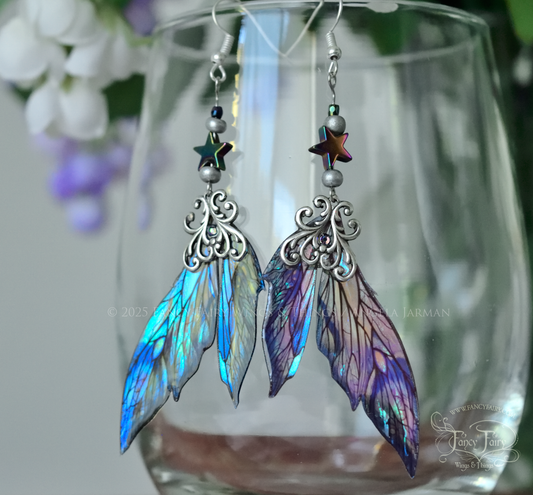 Iridescent fairy wing earrings in blues with silver filigree dangle from the rim of a clear glass