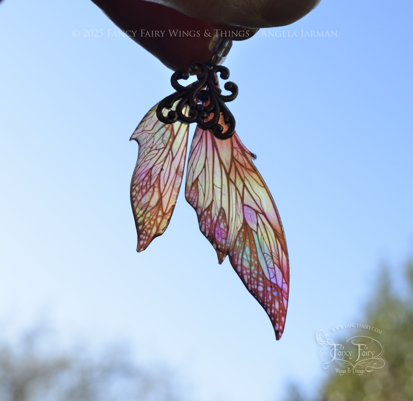 Iridescent fairy wing earrings in earthy colors are back lit with silver filigree dangle from fingers & held up against the blue sky