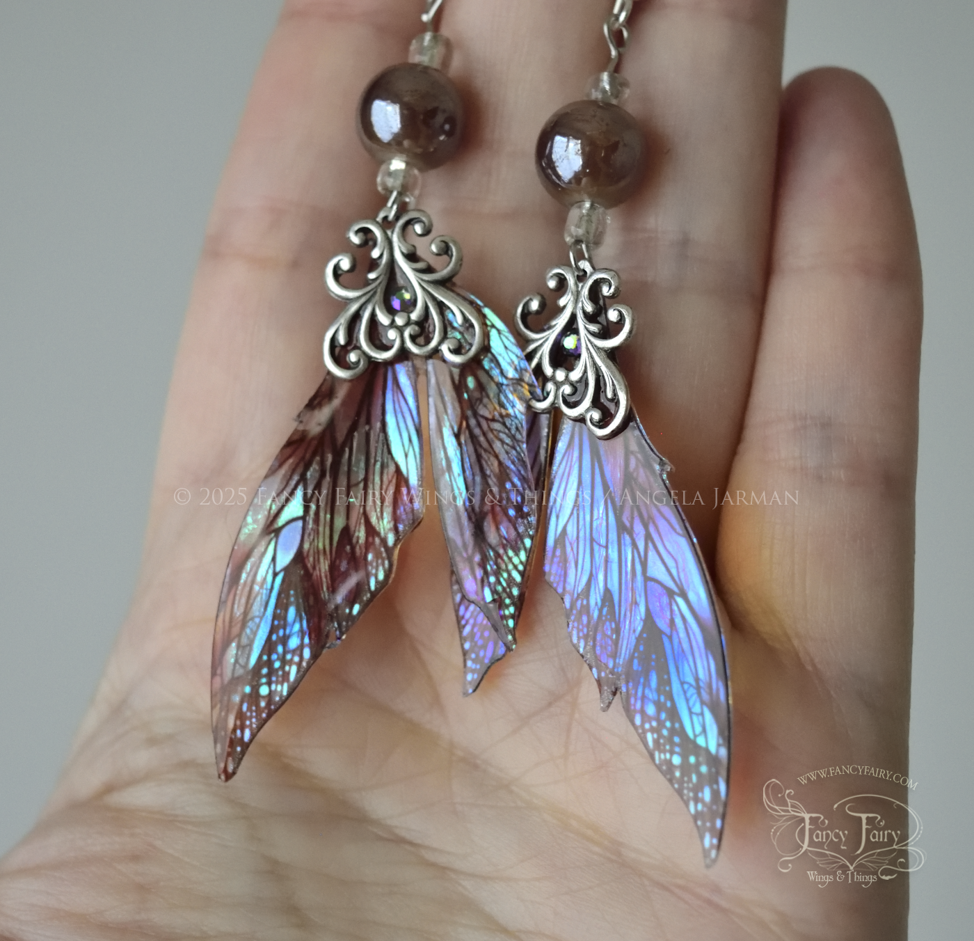 Iridescent fairy wing earrings in earthy colors with silver filigree are held in the palm of a white person's hand