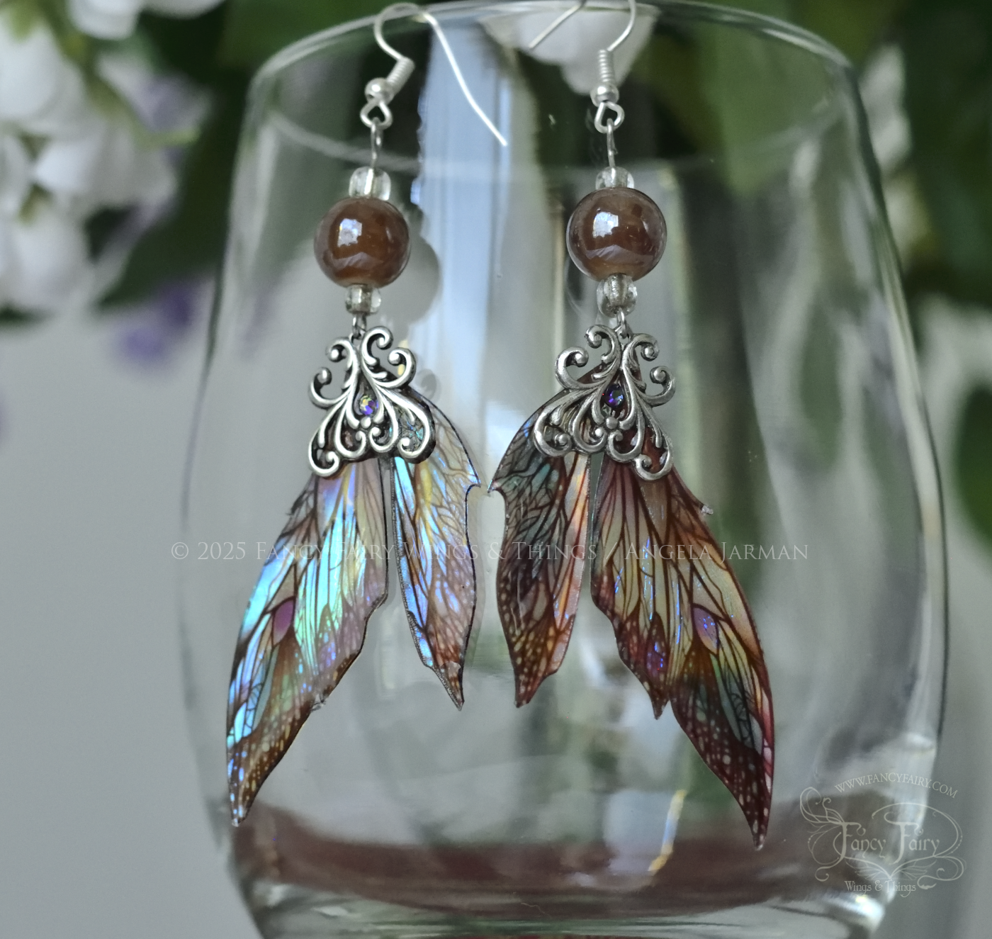 Iridescent fairy wing earrings in earthy colors with silver filigree dangle from the rim of a clear glass