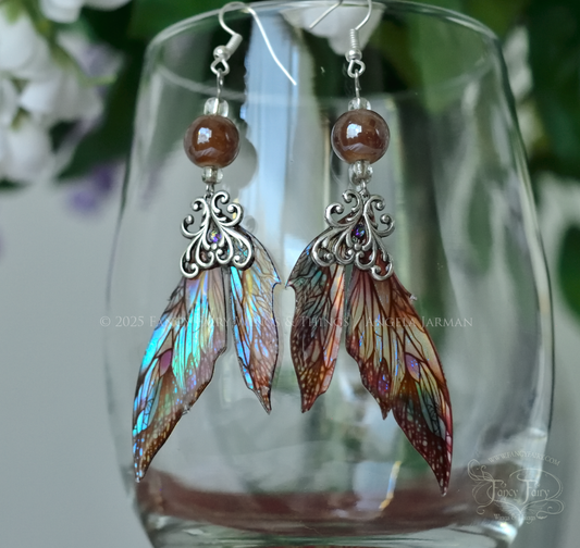 Iridescent fairy wing earrings in earthy colors with silver filigree dangle from the rim of a clear glass