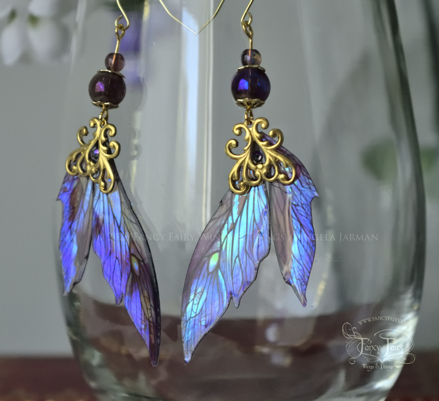 Iridescent fairy wing earrings in purple with brass filigree dangle from the rim of a clear glass