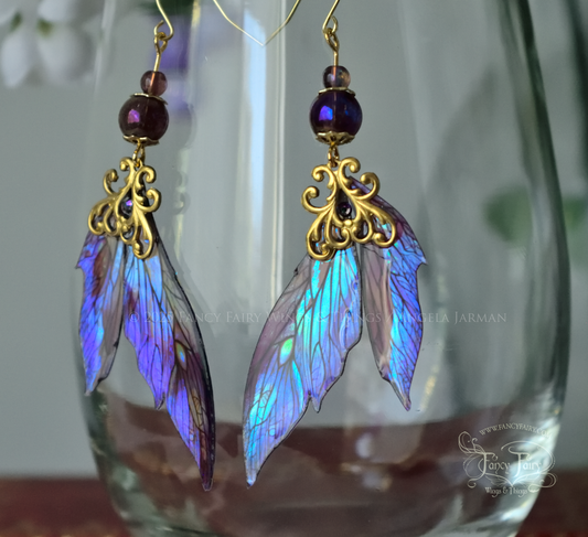 Iridescent fairy wing earrings in purple with brass filigree dangle from the rim of a clear glass