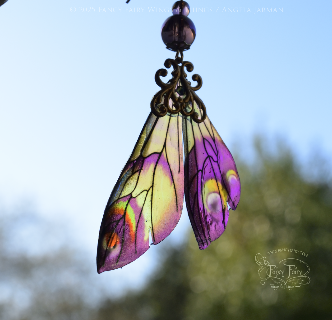 Iridescent fairy wing earrings in plum with green accents and brass filigree held up in front of the sky