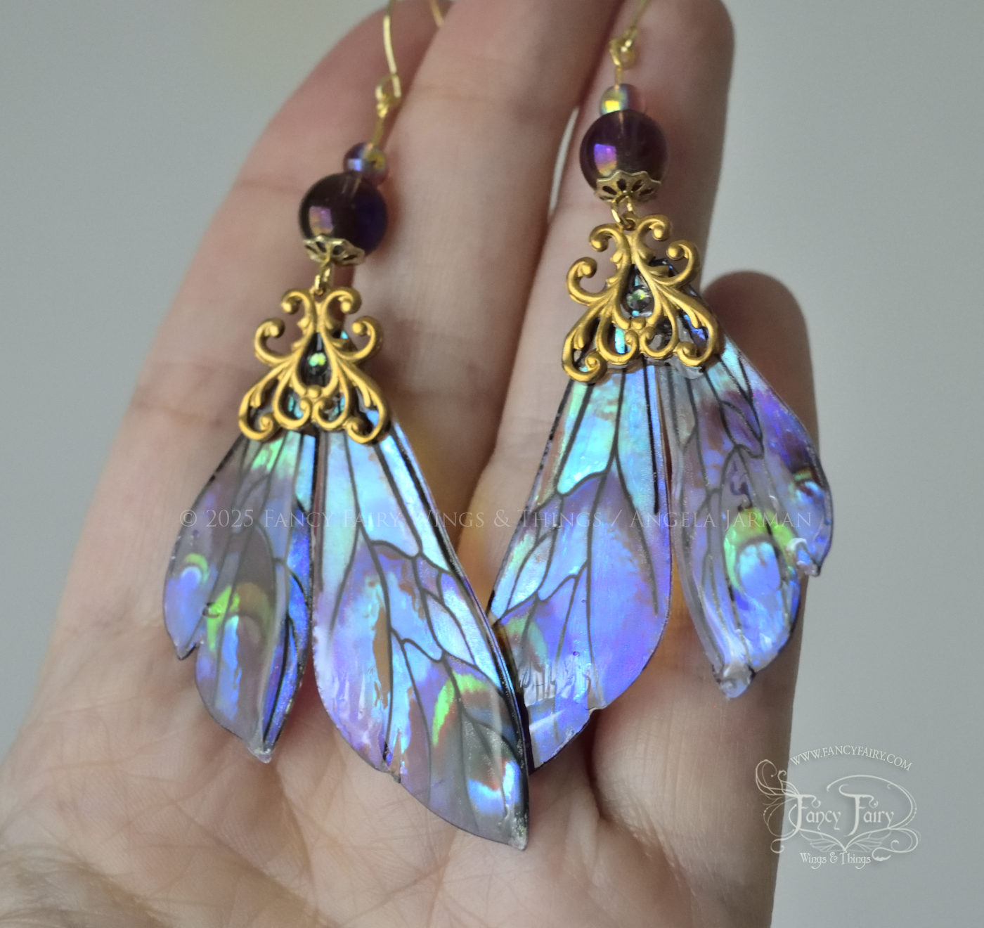 Iridescent fairy wing earrings in plum with green accents and brass filigree held in the palm of a white person's hand