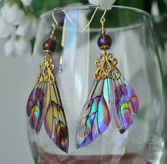 Iridescent fairy wing earrings in plum with green accents and brass filigree dangle from the rim of a clear glass