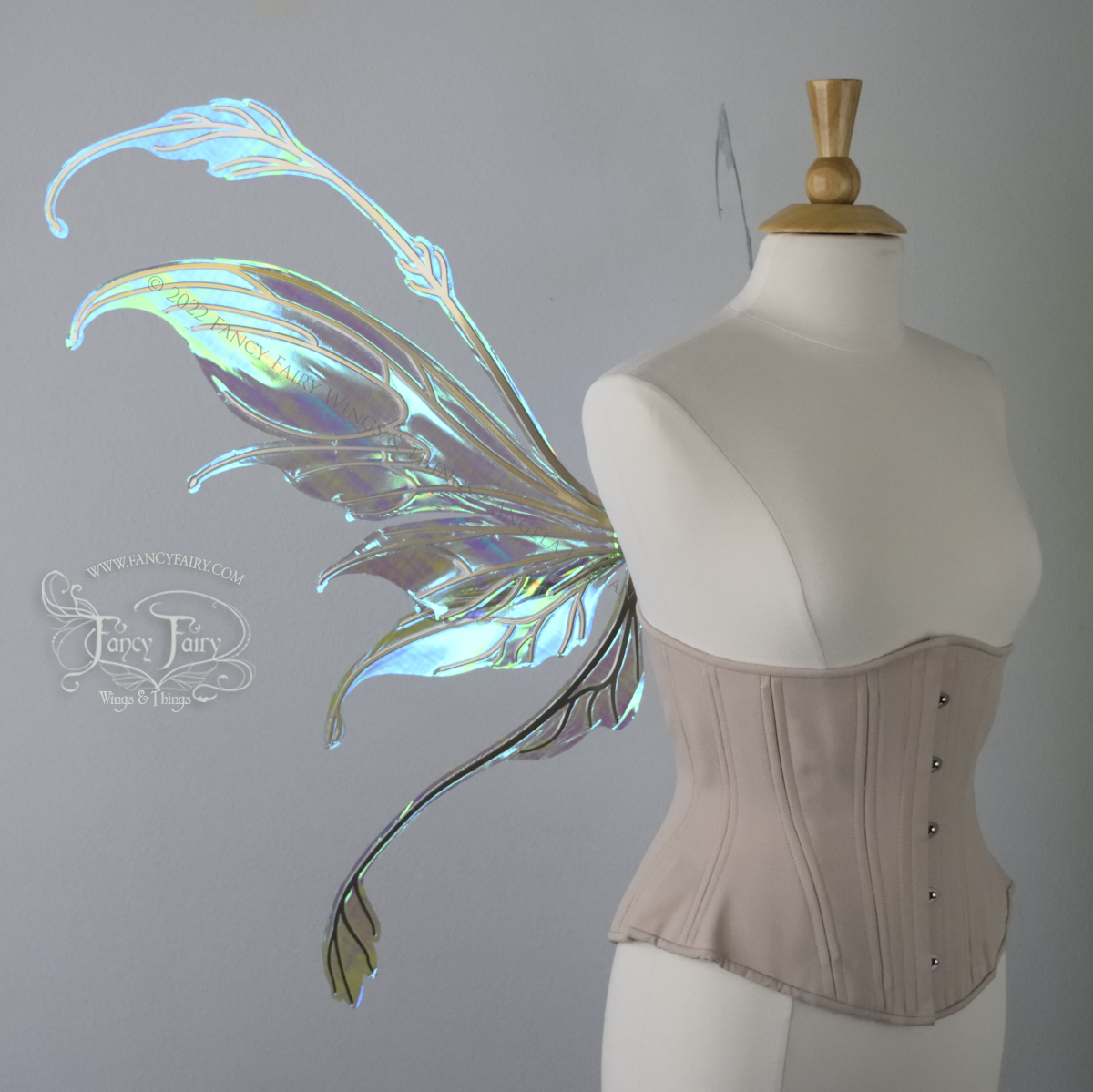 Left side view of an ivory dress form wearing an alabaster underbust corset & large blue/green iridescent fairy wings with gold veins. The upper panels curve downward at top edge with antennae above, bottom panels have a narrow long tail