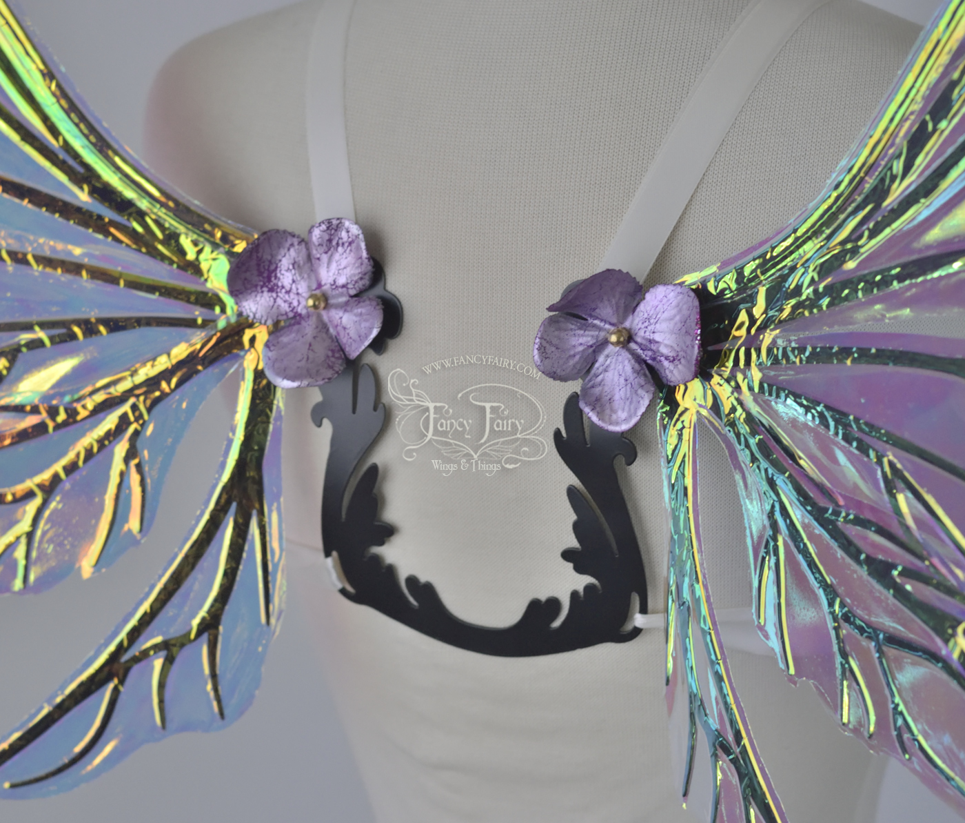 Back closeup view of an ivory dress form wearing an ornate U shaped back brace with iridescent wings sprouting out of the top with shiny silver and violet faux flowers covering the attachment point and secured with a brass acorn cap.