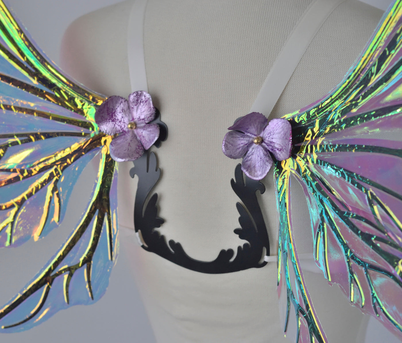 Back closeup view of an ivory dress form wearing an ornate U shaped back brace with iridescent wings sprouting out of the top with shiny silver and violet faux flowers covering the attachment point and secured with a brass acorn cap.