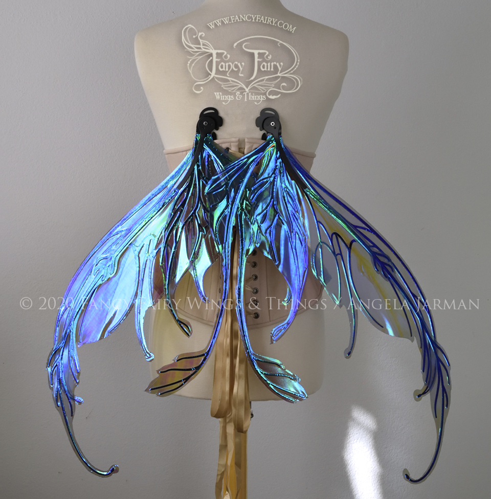 Back view of a dress form wearing an underbust corset & 'Fauna' transparent purple / blue iridescent fairy wings with downward curved tips, antennae & wispy 'tails', with black veining, in resting position