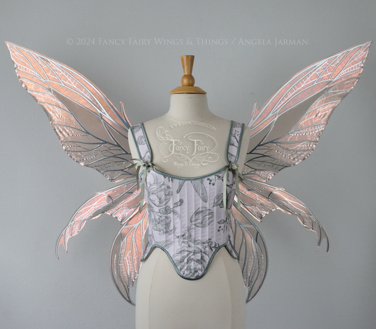 Large rose gold iridescent fairy wings with pointed tips & silver veins worn on a dress form with a white & green corset, front view