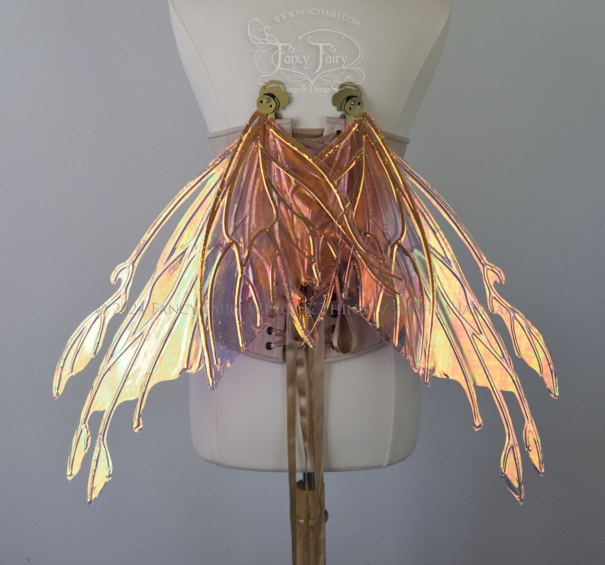Back view of an ivory dress form wearing an alabaster underbust corset & large pink & orange iridescent fairy wings with elongated upper panels with petal like appendages at the tips & antennae, the veins are gold, wings are in resting position