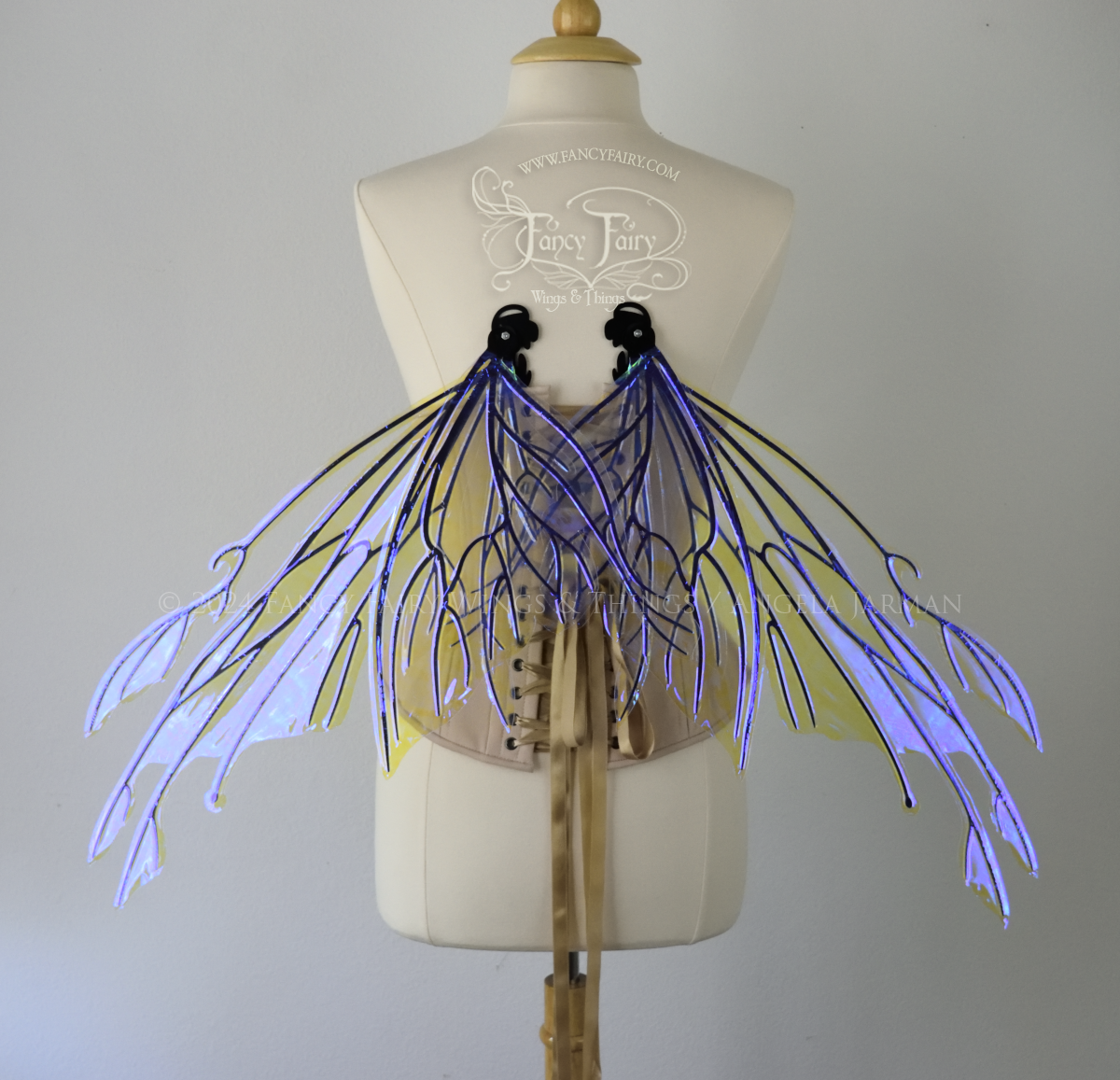Back view of an ivory dress form wearing an alabaster underbust corset & large violet iridescent fairy wings with elongated upper panels with petal like appendages at the tips & antennae, the veins are black, in resting position