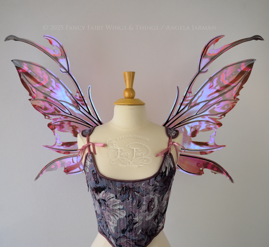 Iridescent fairy wings in mauve, violet, red & black with spikey antennae & tattered edges with holes is worn on a dress form which is also wearing violet & pink floral patterned stays / bodice