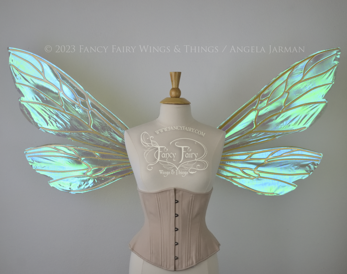 Front view of an ivory dress form wearing an alabaster underbust corset and extra large fairy wings similar to wasp wings, upper and lower panels are both elongated with rounded and slightly pointed tips. They are iridescent green / blue, with gold veins. The background is plain white and my logo and copyright notice are visible.