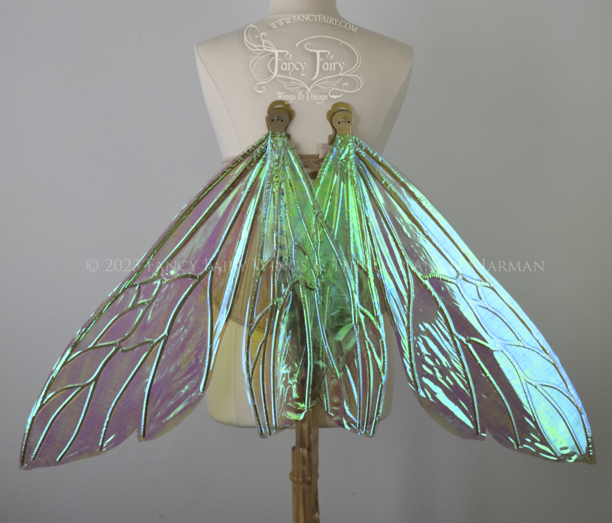 Front view of an ivory dress form wearing an alabaster underbust corset and extra large fairy wings similar to wasp wings, upper and lower panels are both elongated with rounded and slightly pointed tips. They are iridescent green / blue, with gold veins. Wings are in resting position. The background is plain white and my logo and copyright notice are visible.
