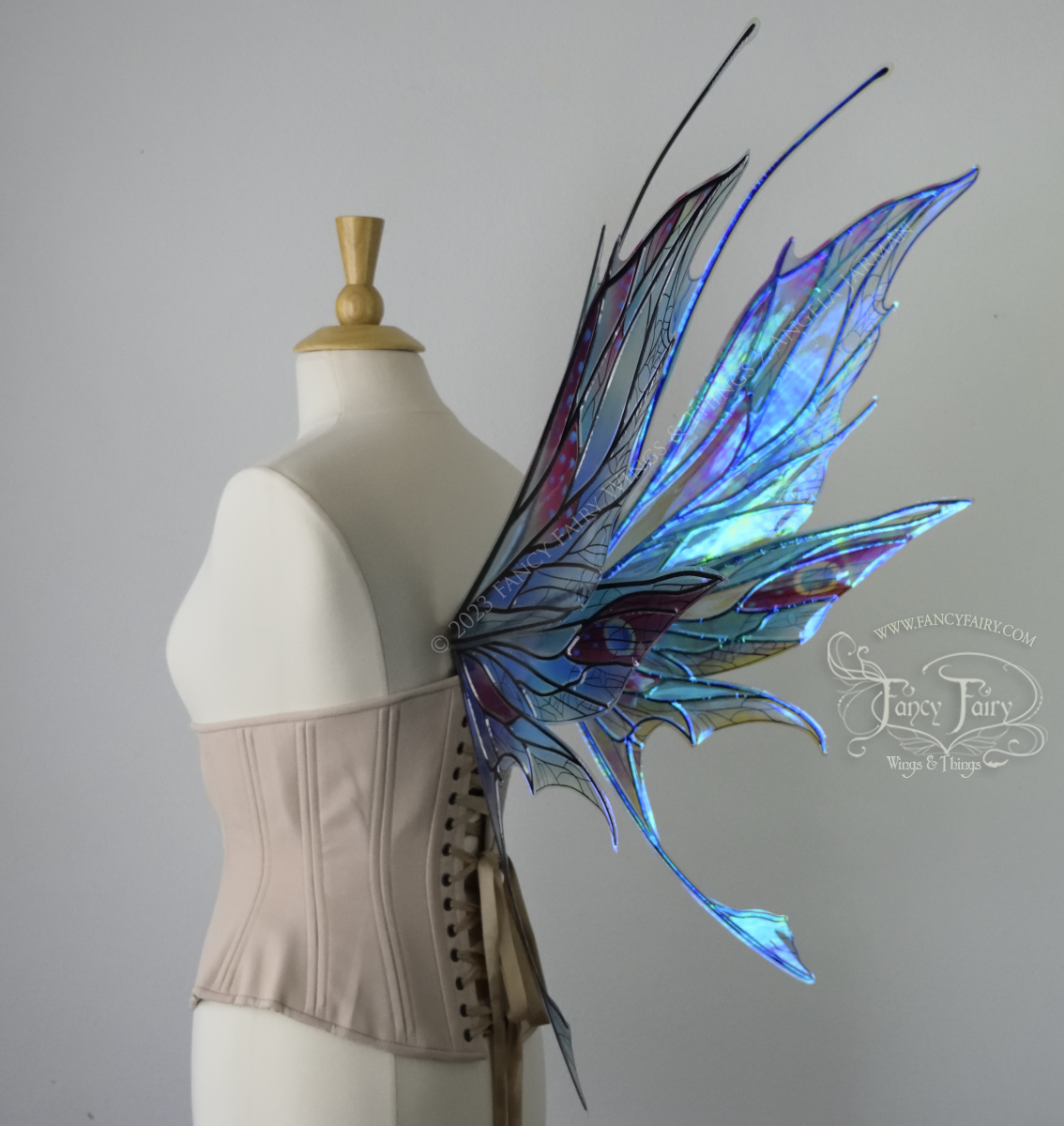 Back 3/4 view of an ivory dress form wearing an alabaster underbust corset and large multicolor iridescent fairy wings featuring antennae along the top. Upper panels come to a point as do the middle panels, bottom panels have tails. Spikey, detailed veins