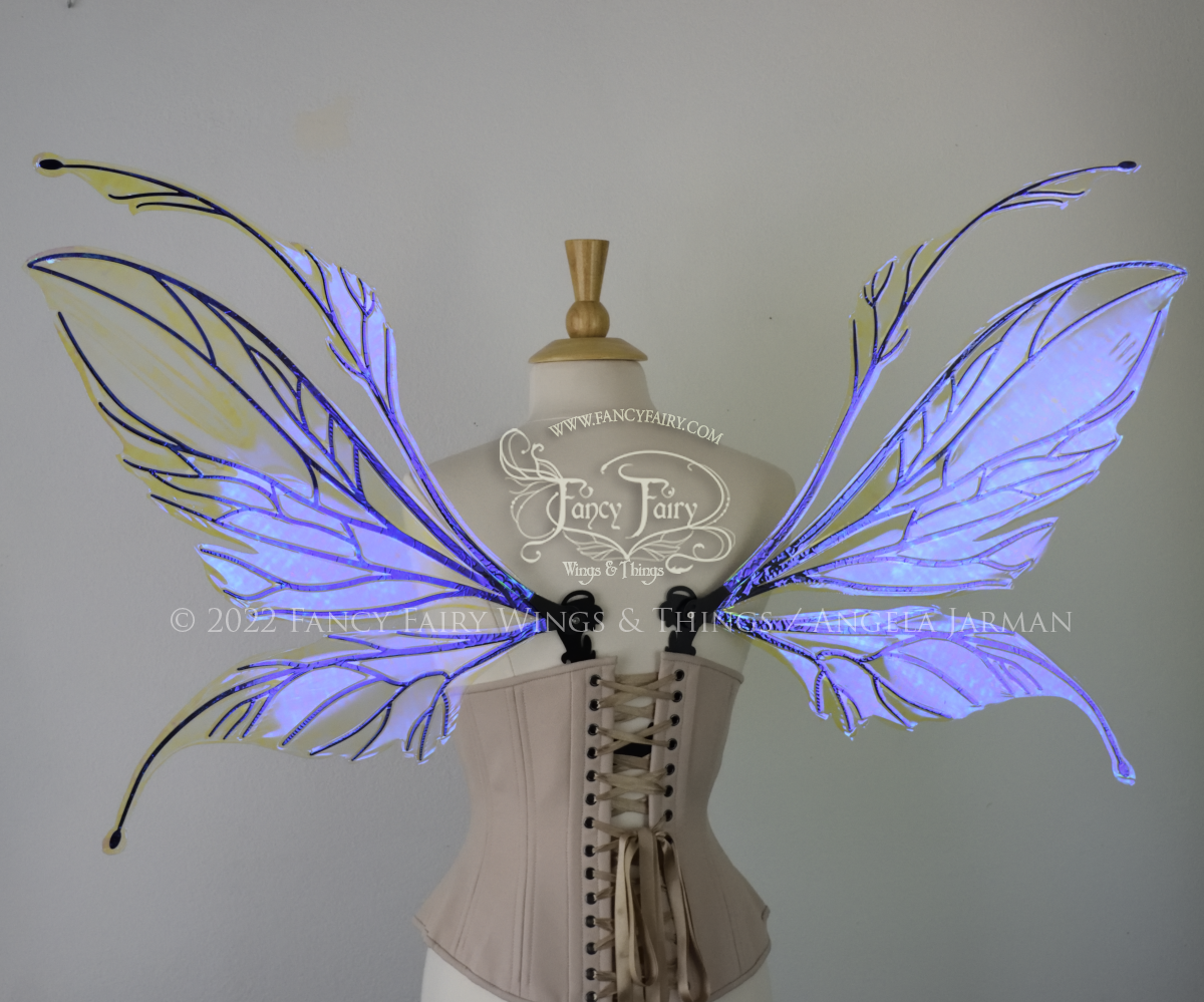 Back view of an dress form wearing an underbust corset and large iridescent violet fairy wings featuring antennae along the top. The upper panels are elongated with semi-pointed tips, the lower panels are smaller with slender tips that curve downward. The veins are black