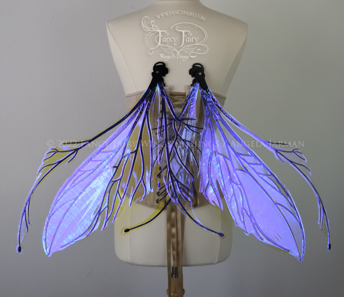 Back view of an dress form wearing an underbust corset and large iridescent violet fairy wings featuring antennae along the top. The upper panels are elongated with semi-pointed tips, the lower panels are smaller with slender tips that curve downward. The veins are black, & the wings are in resting position