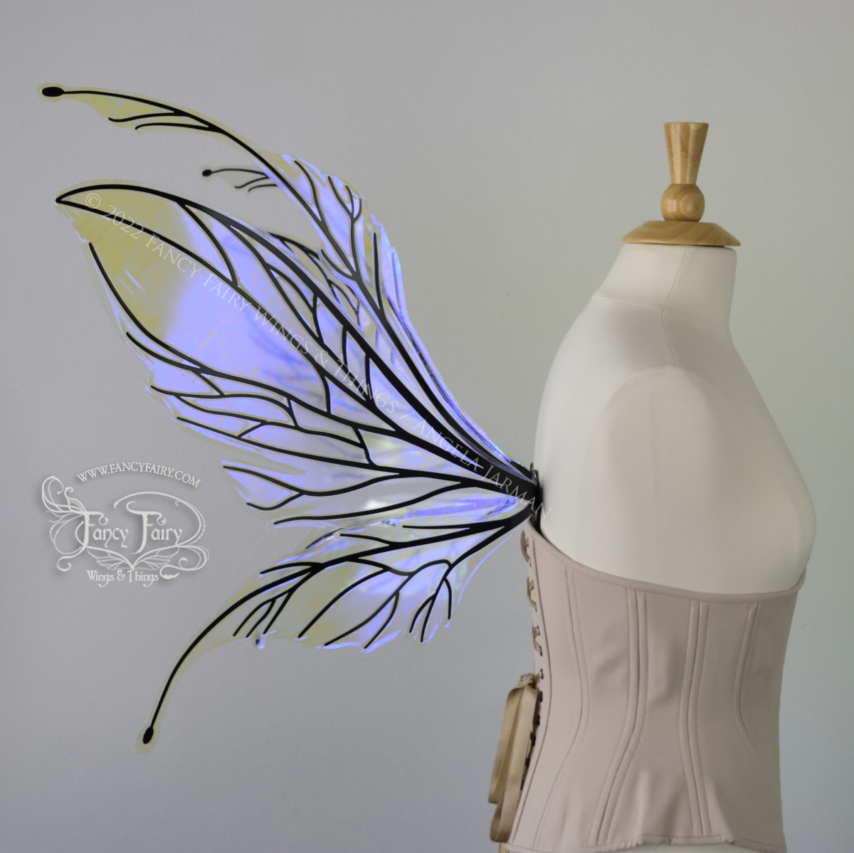 Left side view of an dress form wearing an underbust corset and large iridescent violet fairy wings featuring antennae along the top. The upper panels are elongated with semi-pointed tips, the lower panels are smaller with slender tips that curve downward. The veins are black