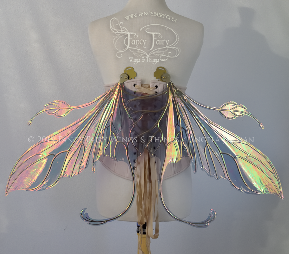 Back view of an ivory dress form wearing an alabaster underbust corset & large orange/green iridescent fairy wings with elongated upper panels & antennae with bottom panels that have a tail curving upwards, gold veins, in resting position