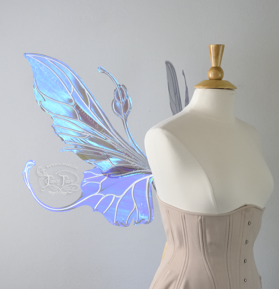 Left side view of an ivory dress form wearing an alabaster underbust corset & large purple/blue iridescent fairy wings with elongated upper panels & antennae with bottom panels that have a tail curving upwards, silver veins