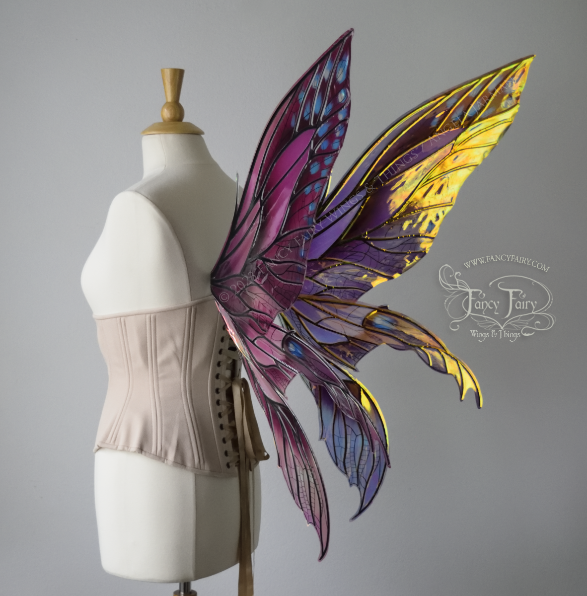 Back 3/4 side view of large burgundy and peach pink iridescent fairy wings with 3 panels on each side, pointed tips, lots of vein detail, worn on a dress form.