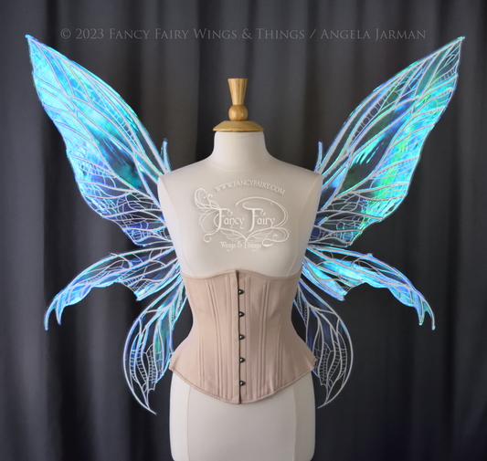Front view of an ivory dress form wearing an underbust corset and large blue/teal/violet iridescent fairy wings. Upper panels are elongated with pointed tips, 2 lower panels curve downward, detailed white veins, grey background 