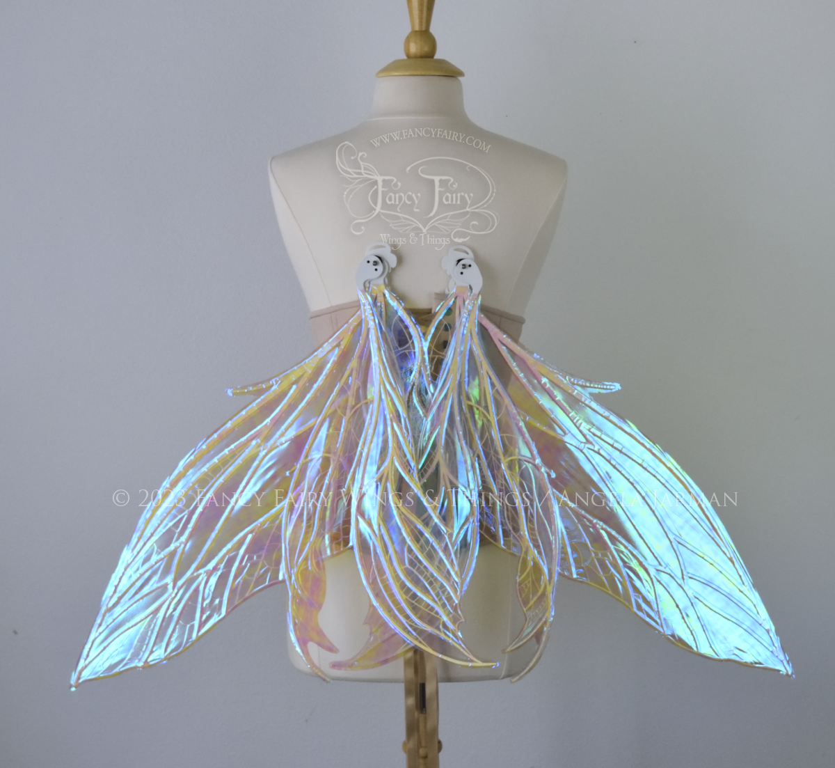 Back view of an ivory dress form wearing an underbust corset and large blue/teal/violet iridescent fairy wings, in the resting position angled downward. Upper panels are elongated with pointed tips, 2 lower panels curve downward, detailed white veins 