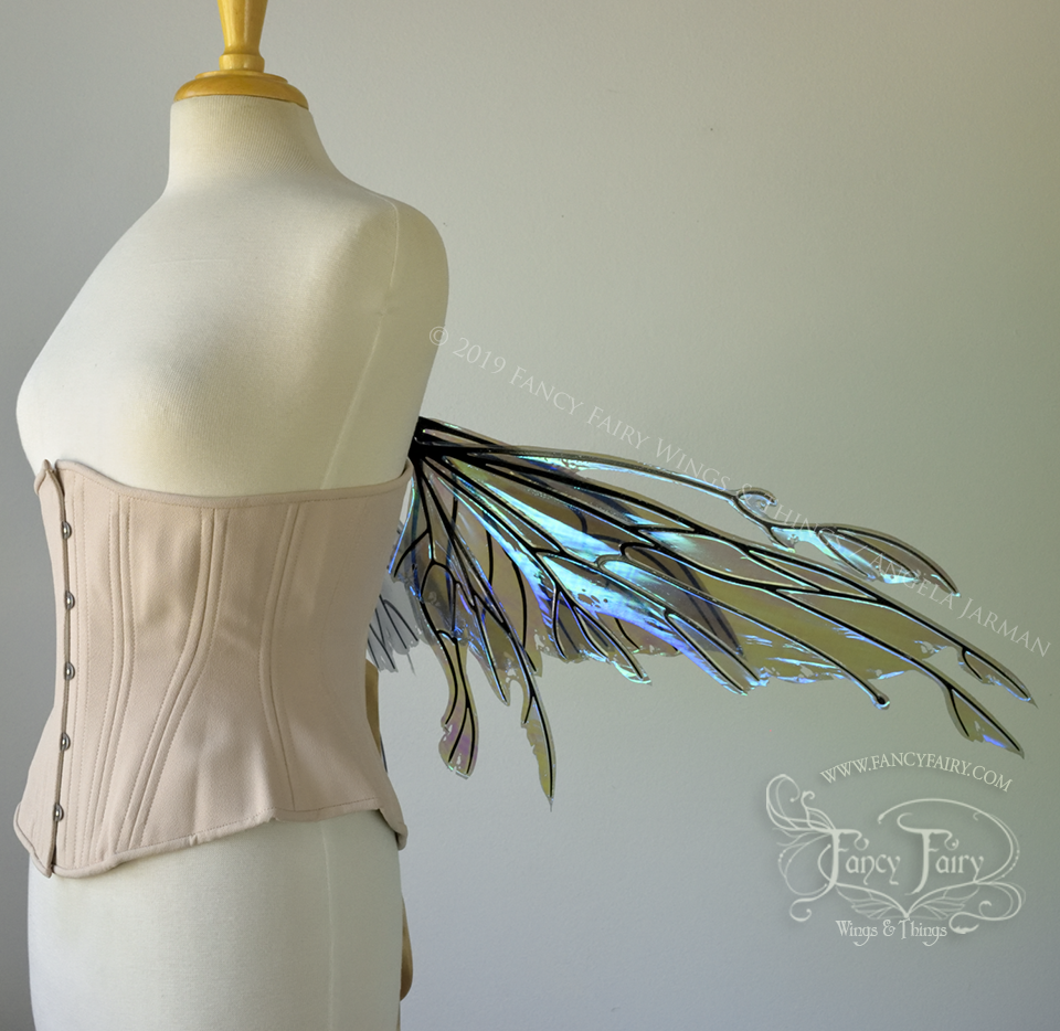 Right side view of an ivory dress form wearing an alabaster underbust corset & purple / blue iridescent fairy wings with elongated upper panels with petal like appendages at the tips & antennae, the veins are black. Wings are lowered in resting position